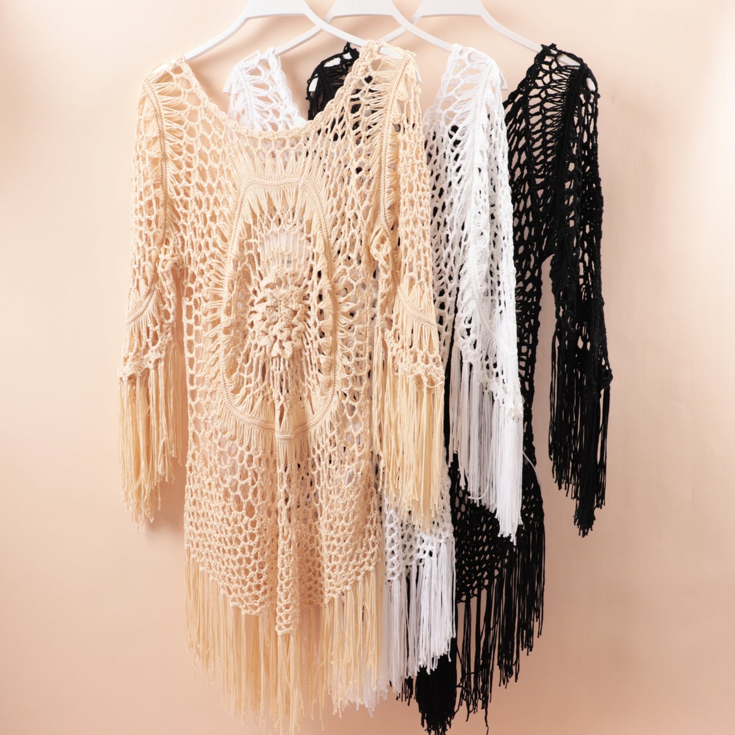 Three Macrame fringe garments in beige, white, and black on hangers against a beige background.