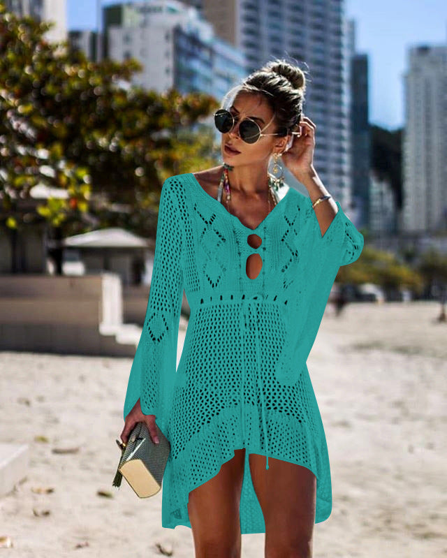 Woman wearing a teal crochet dress on a beach with cityscape in the background