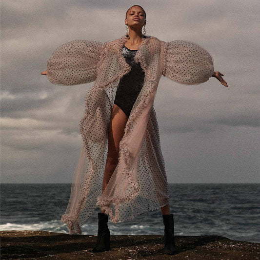 Women wearing a long, sheer polka dot cover-up over a black swimsuit by the ocean.